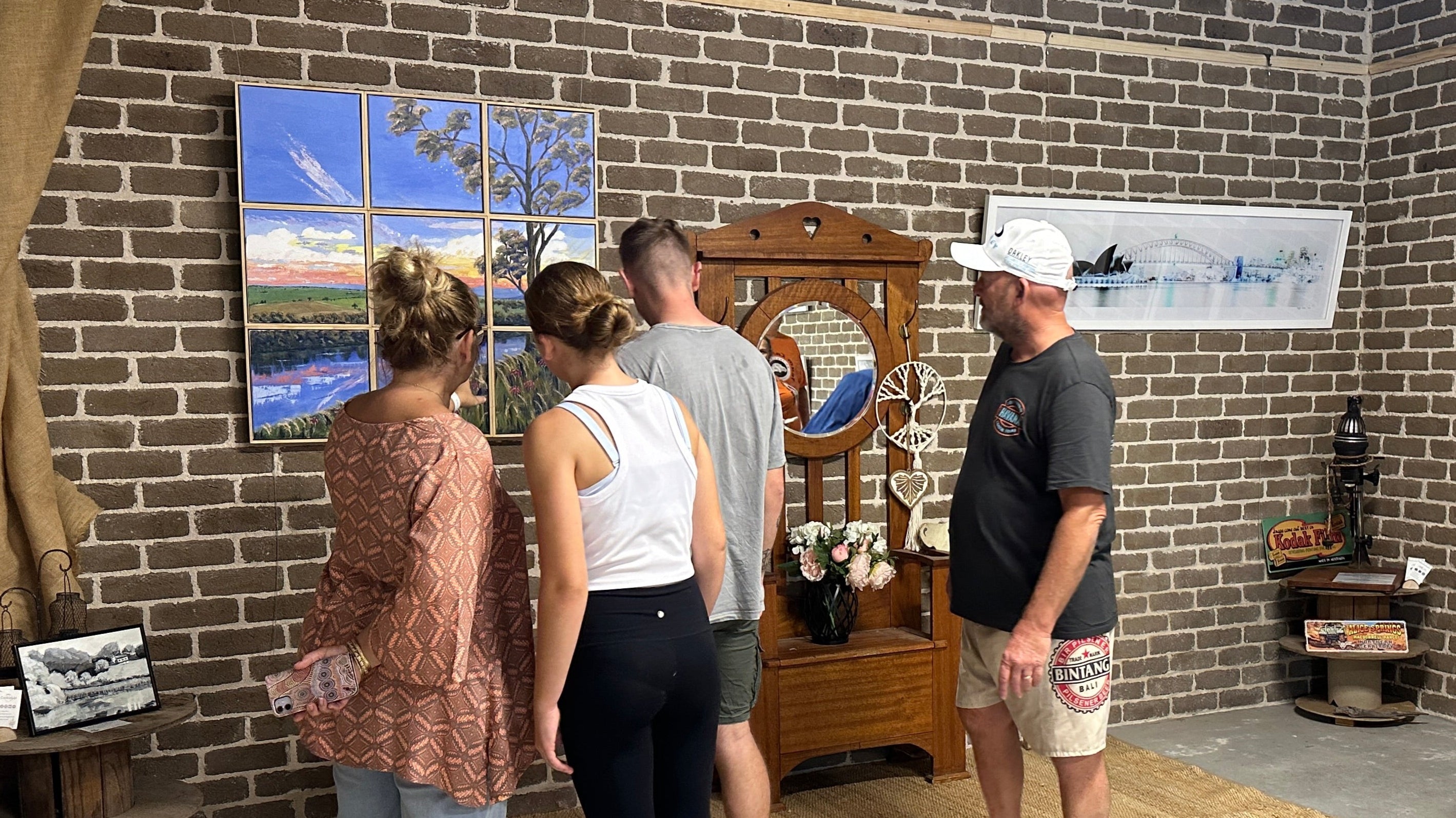 Group of people in a room with brick walls, looking at various items including a clock and framed pictures.