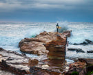 Person with a surfboard standing on a rocky outcrop with large waves crashing around