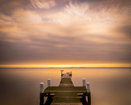 Wooden dock extending into a calm body of water with a dramatic sky during sunset.