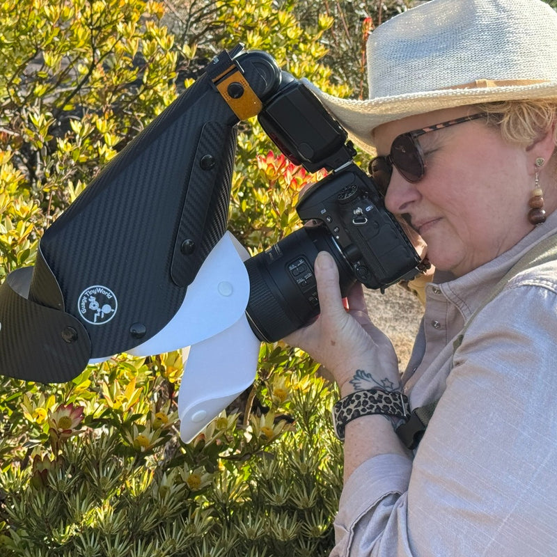 Person holding a camera with a macro hood and flash attachment against a bush background