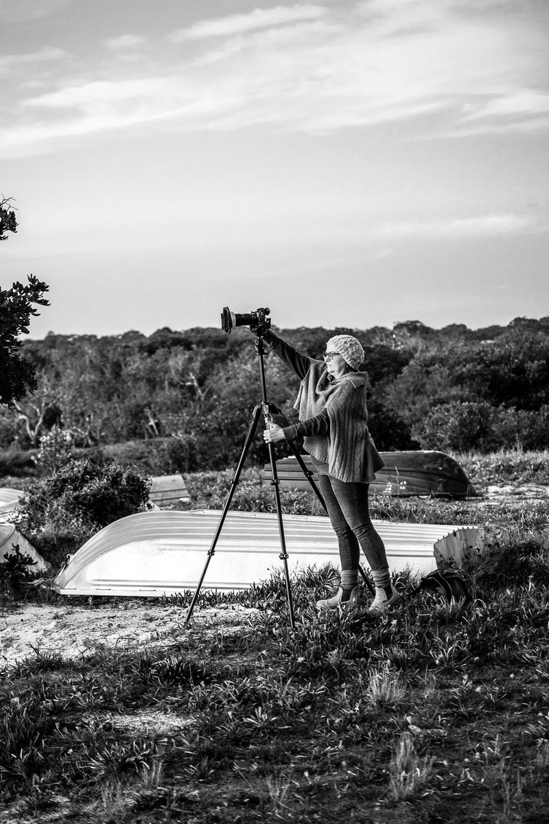 Black and white image of a person with camera on a tripod next to an upturned boat in a coastal setting