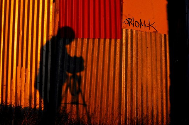 Shadow of a person holding a camera on tripod against a red orange corrugated metal wall with graffiti