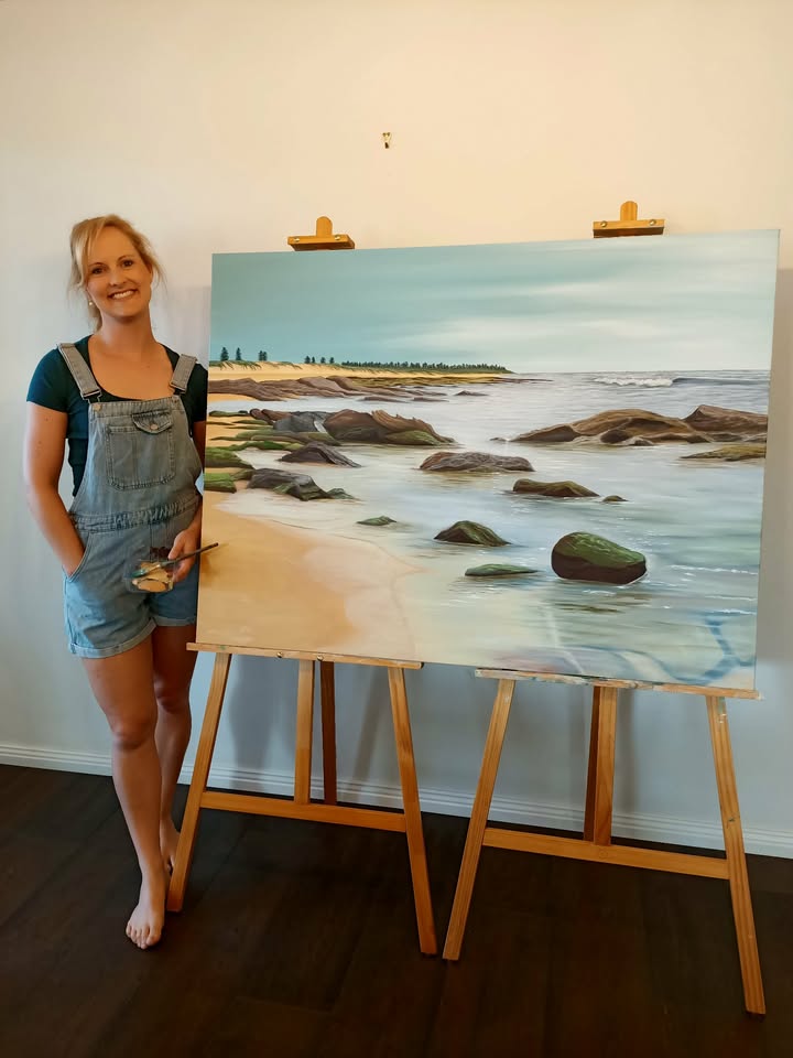 Artist standing next to a painting of a large coastal scene resting across two easels.