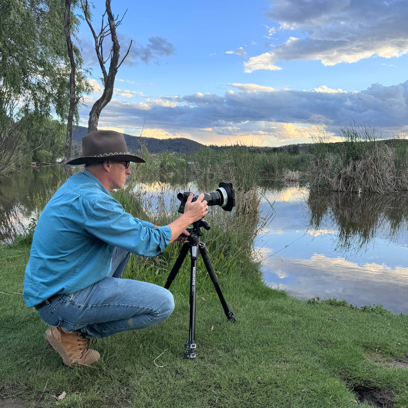 Man wearing a cowboy hat taking a photograph of a scenic lake with a camera on a tripod.