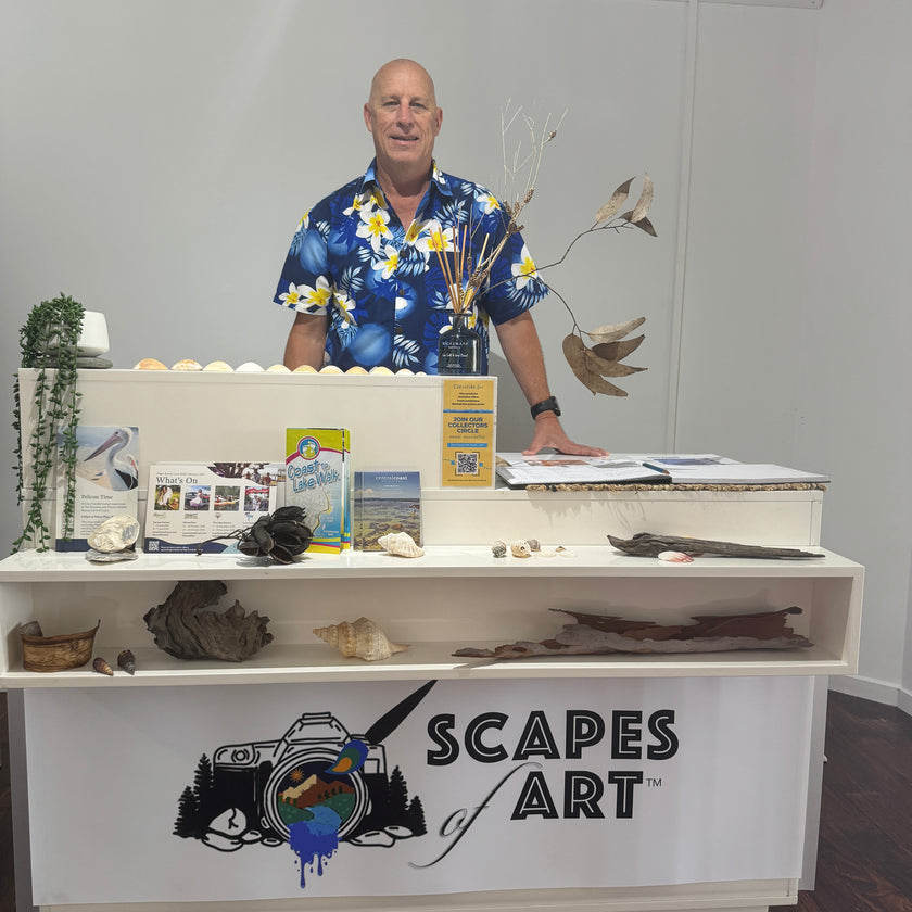 Man standing behind a display counter with 'Scapes of Art' branding, featuring various coastal decorations