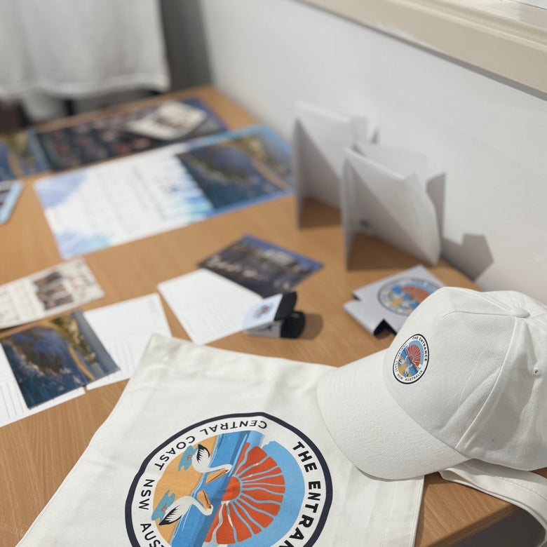 White tote bag and cap with a circular logo of The Entrance Central Coast NSW and two pelicans on a wooden table.