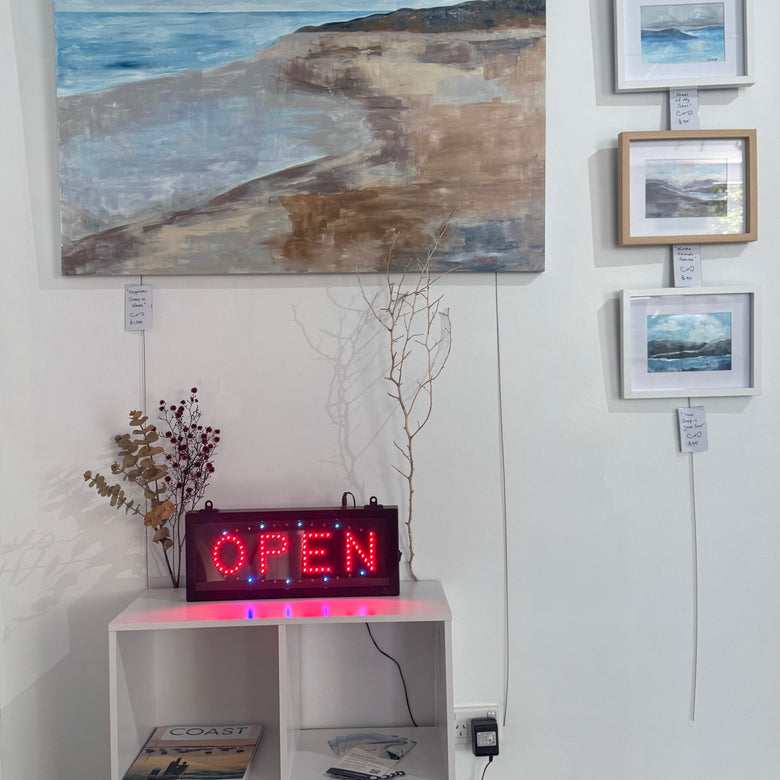 Art gallery with framed paintings on the wall and a 'OPEN' sign on a shelf.