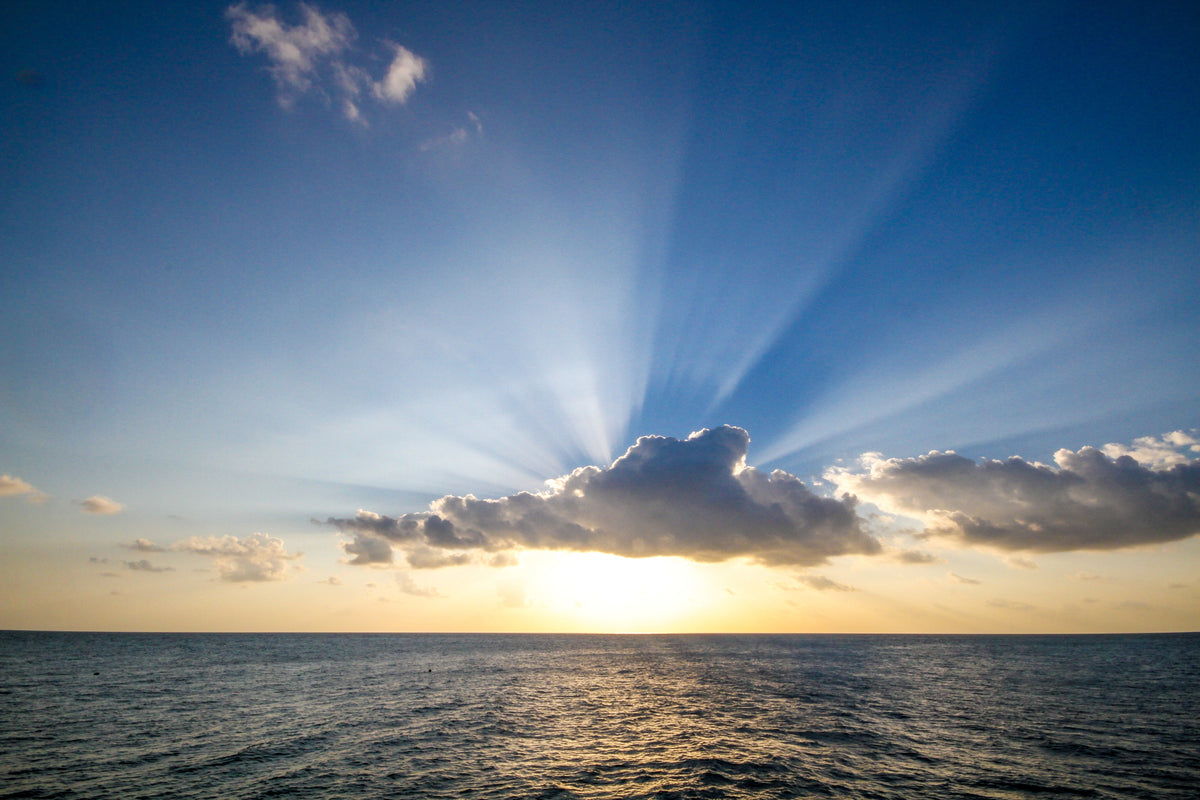 Sun rising through clouds with crepuscular rays over a calm ocean with blue sky